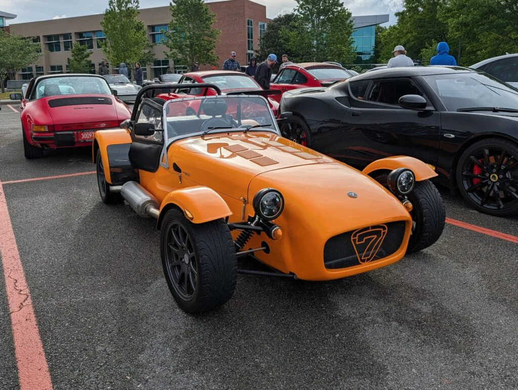 Orange Caterham 7 parked in a lot with several other cars in the background, including a red Porsche and a black Lotus.