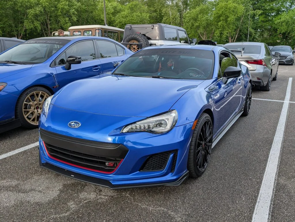 Blue Subaru BRZ with aftermarket modifications parked in a lot next to other vehicles and greenery in the background.