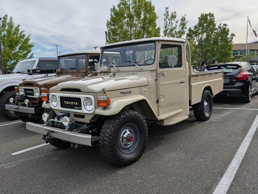 Tan Toyota Land Cruiser pickup truck parked among other vehicles in a lot, surrounded by greenery and event attendees.