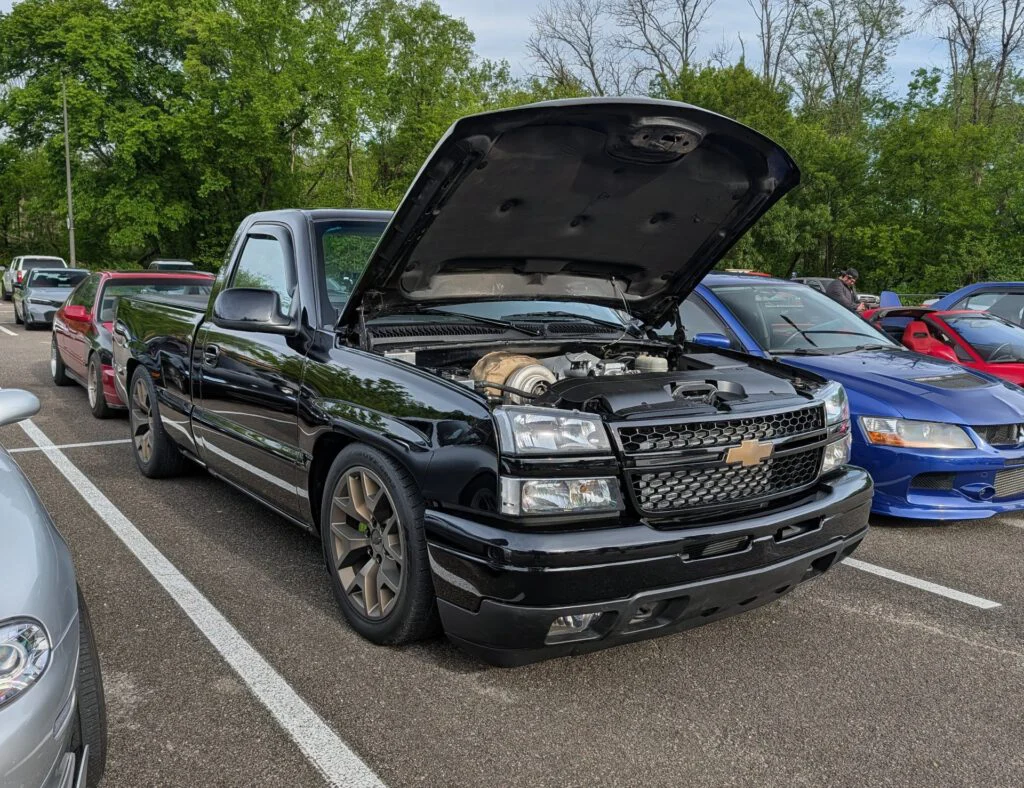 Black Chevrolet pickup truck with hood open, showcasing engine; parked among other cars in a lot with green trees in the back