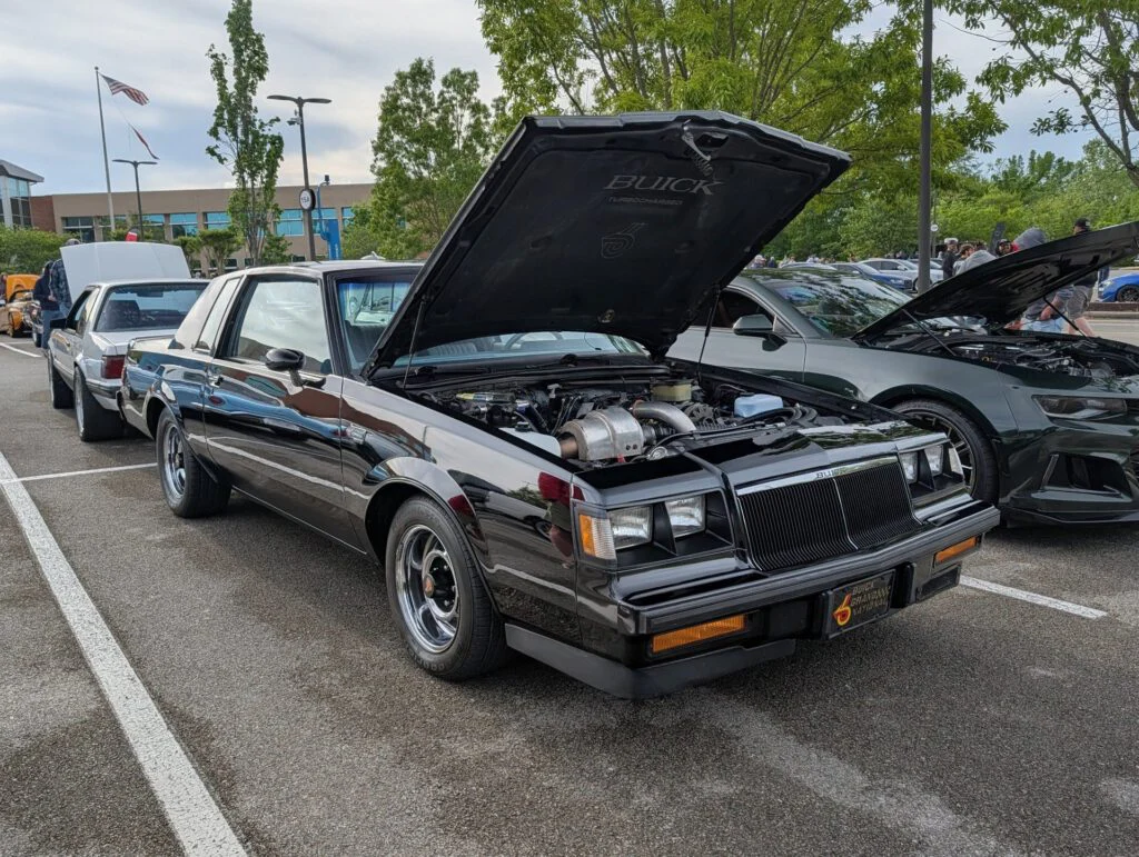 Black Buick with an open hood parked in a lot, showing modifications under the hood. Other vehicles are visible in the backgr