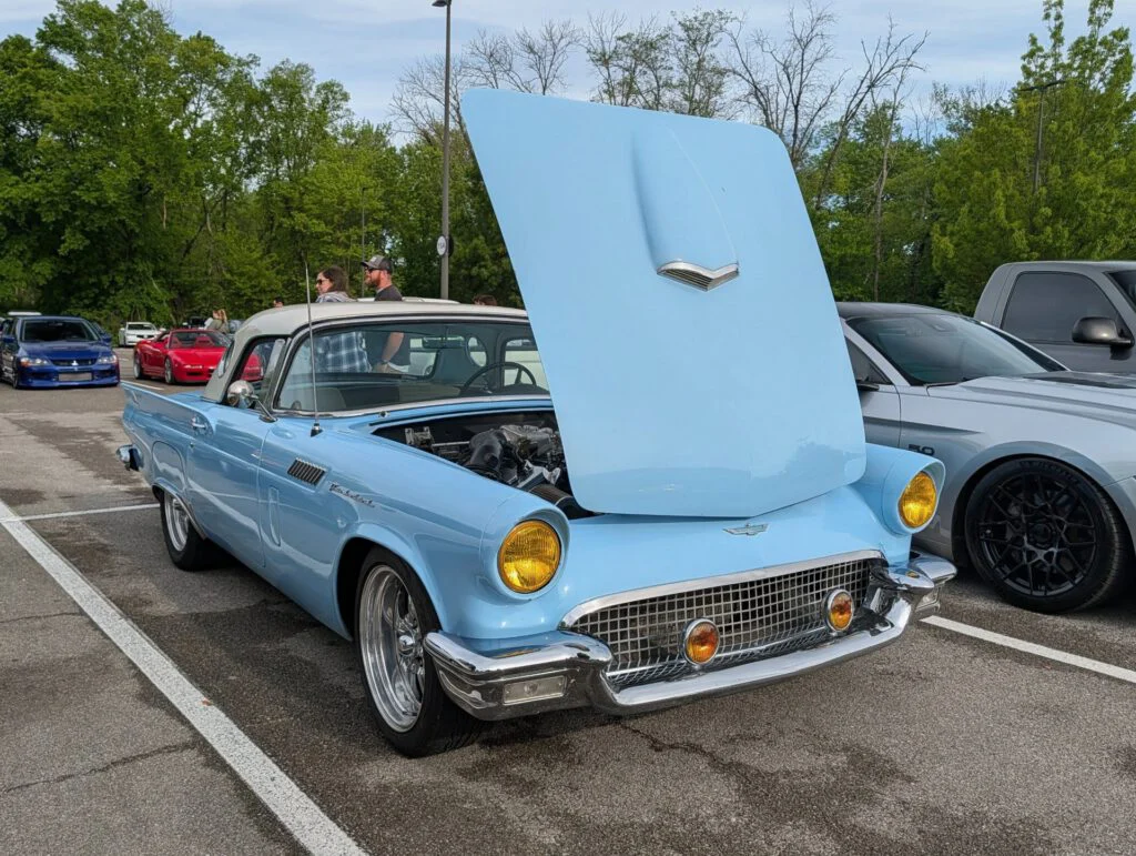 Light blue convertible with an open hood parked in a lot, surrounded by other vehicles and greenery in the background.