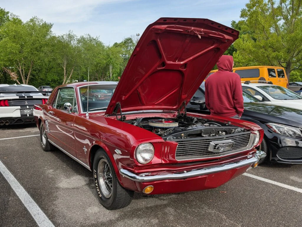 Red Ford Mustang with the hood raised, parked in a lot with various cars in the background and a person standing nearby.