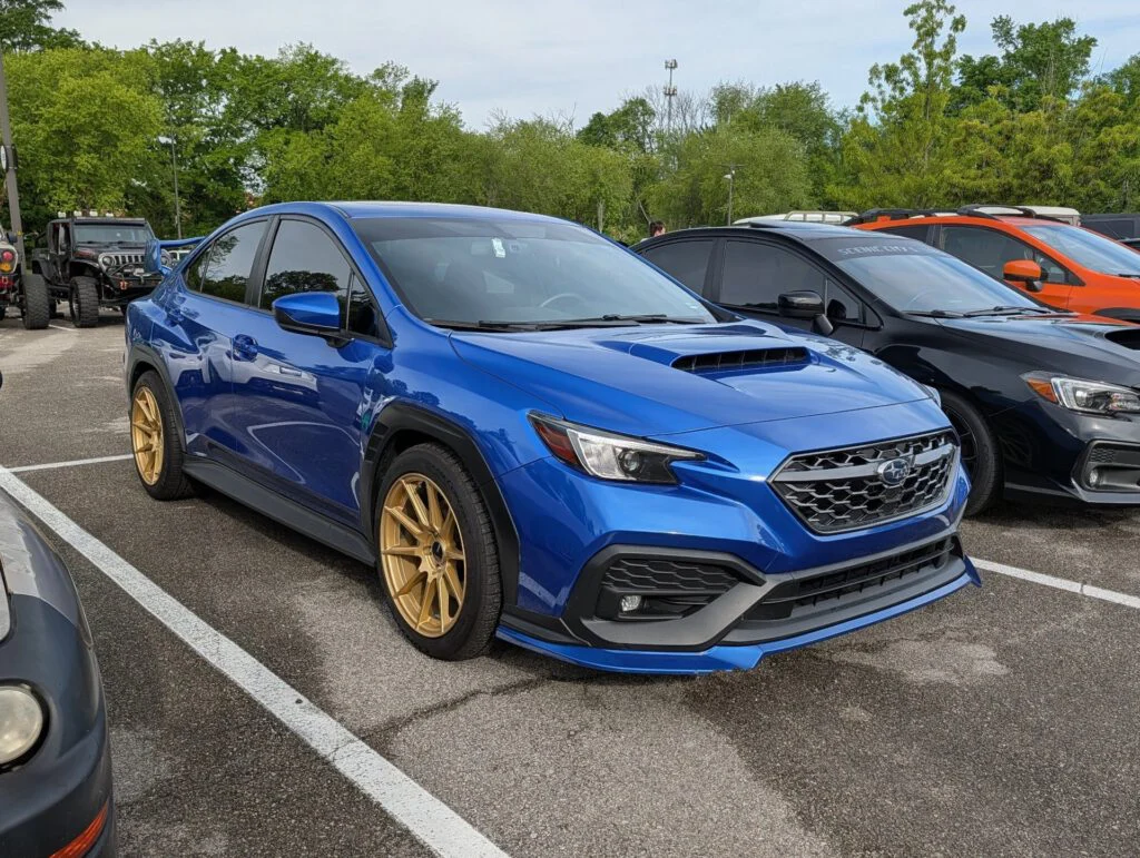 Blue Subaru vehicle with gold wheels parked in a lot among other cars and trees in the background.