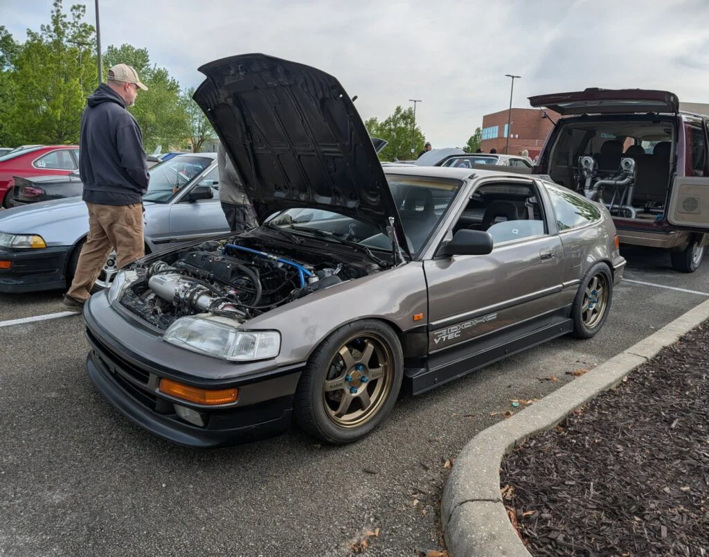 A gray Honda Civic with the hood open, showcasing the engine and modifications, parked in a lot with other cars in the backgr