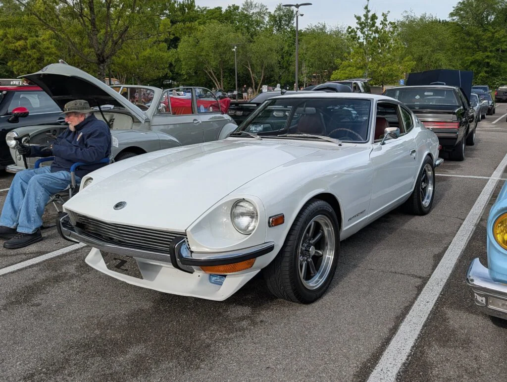 White Datsun 240Z parked in a lot with a man sitting in a chair nearby and other vehicles in the background.
