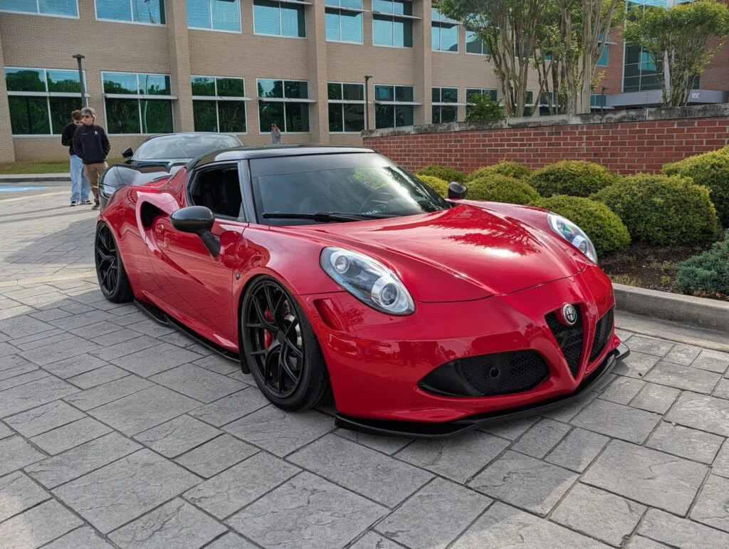 Red Alfa Romeo 4C parked on a paved area with bushes and buildings in the background.