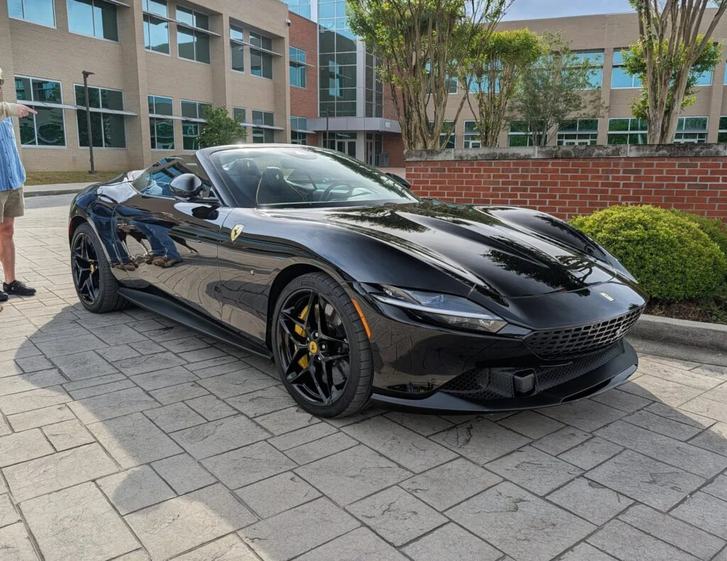 Black convertible sports car parked on paved surface in front of a building with glass windows and greenery.