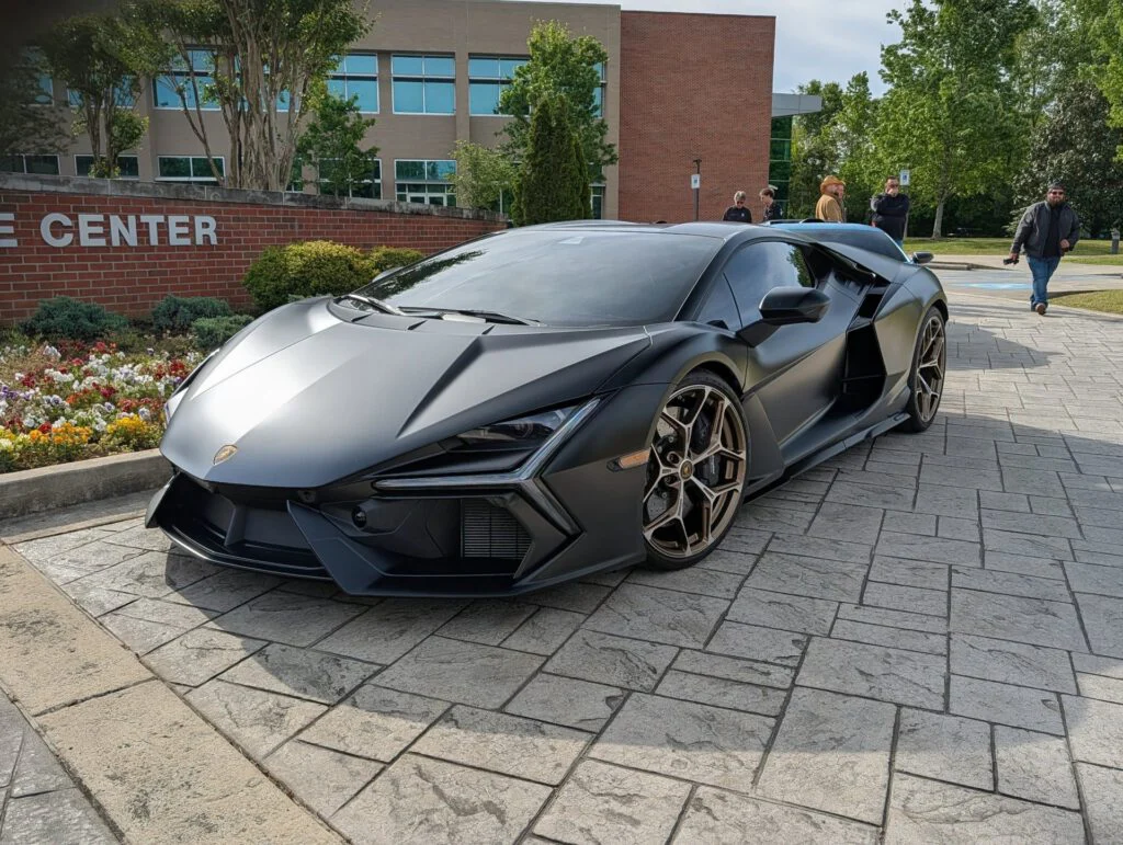 Lamborghini Revuelto in matte black parked near a flower bed with attendees in the background.