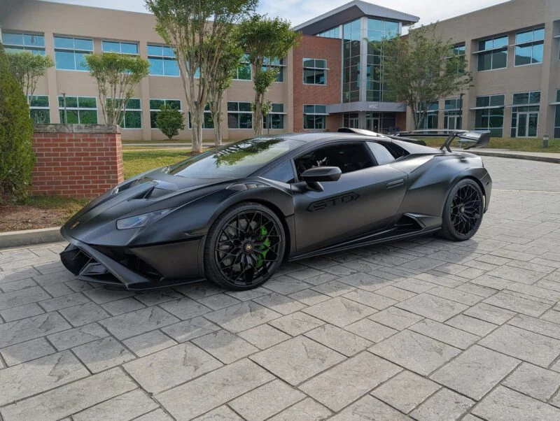 A matte black Lamborghini Huracán STO parked on a paved area in front of a building, with green accents on the wheels.