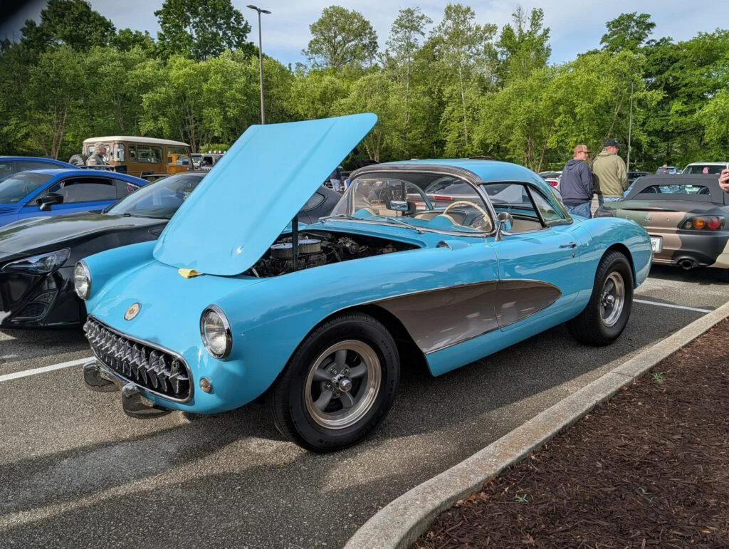 A blue Chevrolet Corvette with its hood raised, parked at a car event with other vehicles and attendees in the background.