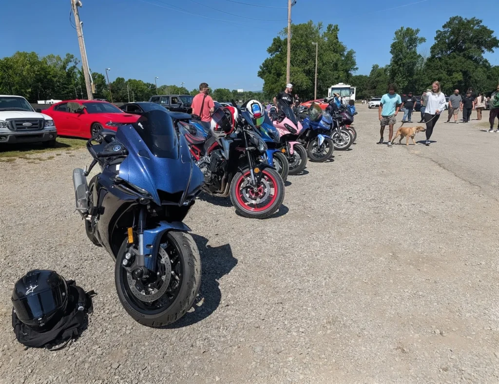 Several motorcycles lined up in a parking area during an event, with attendees and vehicles in the background.