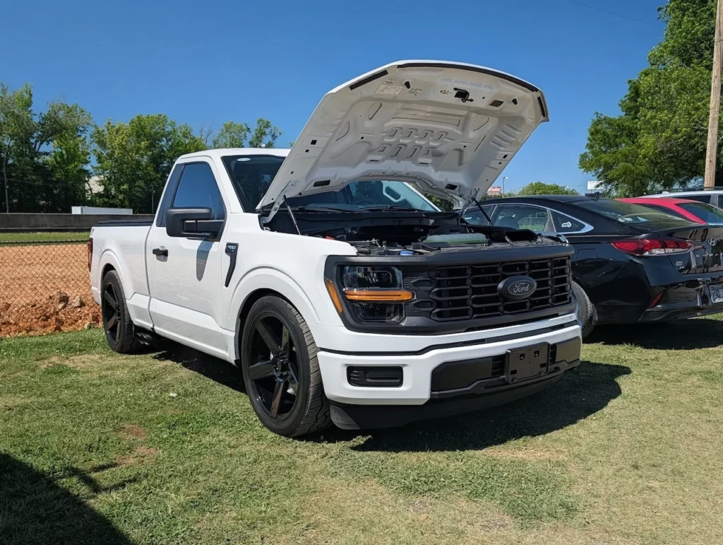 White Ford F-150 with the hood open on a grassy area at an outdoor event.
