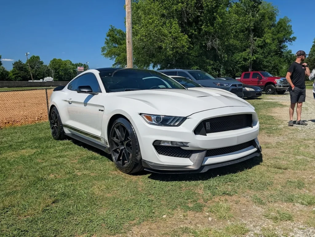White Ford Mustang Shelby GT350 parked on grass with trees in the background.