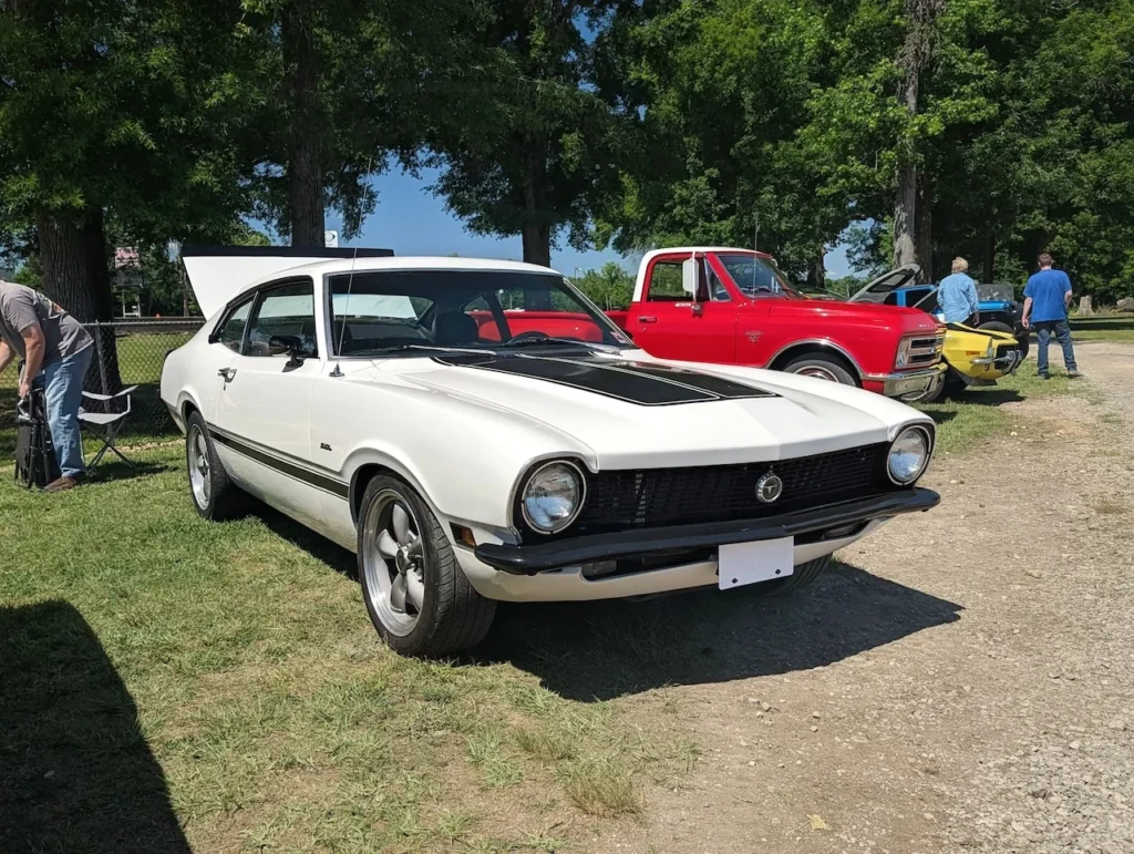 White Ford Maverick parked on grass at outdoor car event with other vehicles.