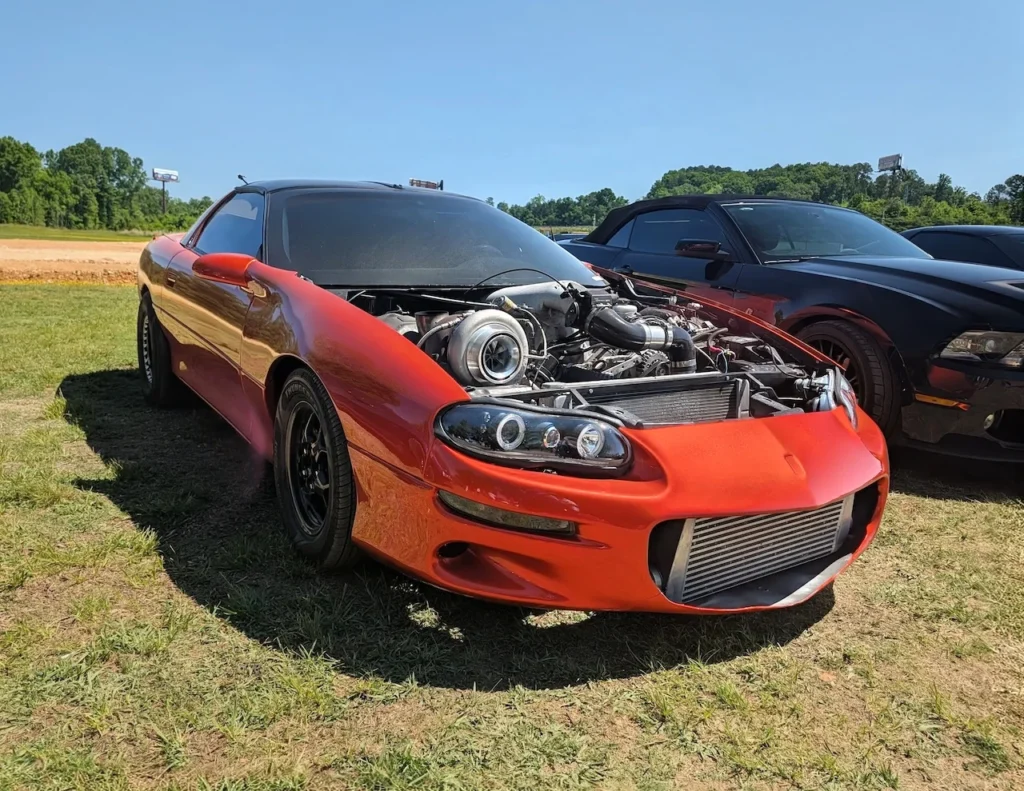 A red Chevrolet Camaro with an open engine bay and custom modifications on a grassy area, with another car in the background.