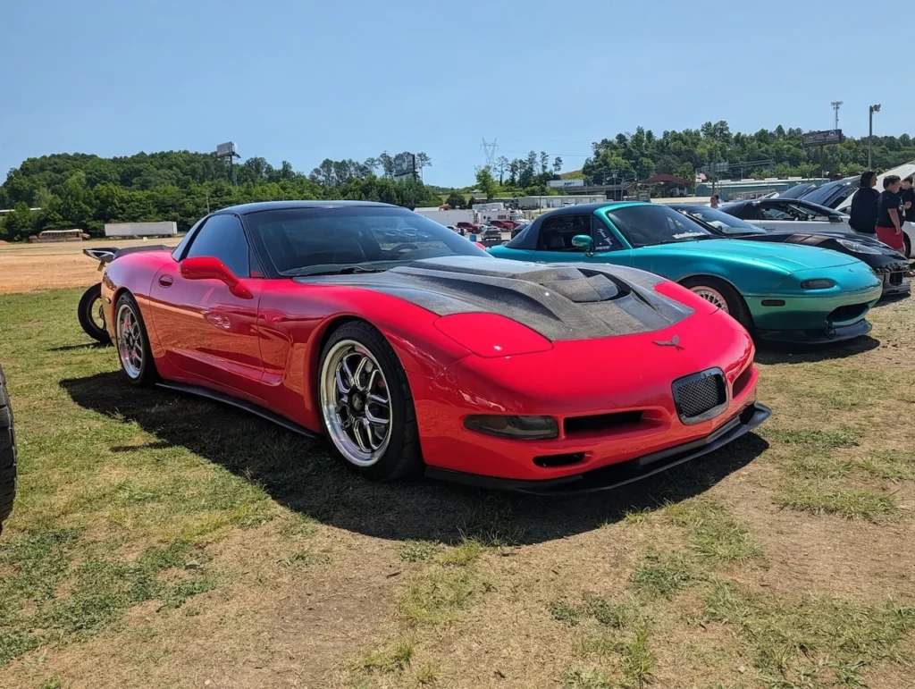 Red sports car with modified hood at a car show field.