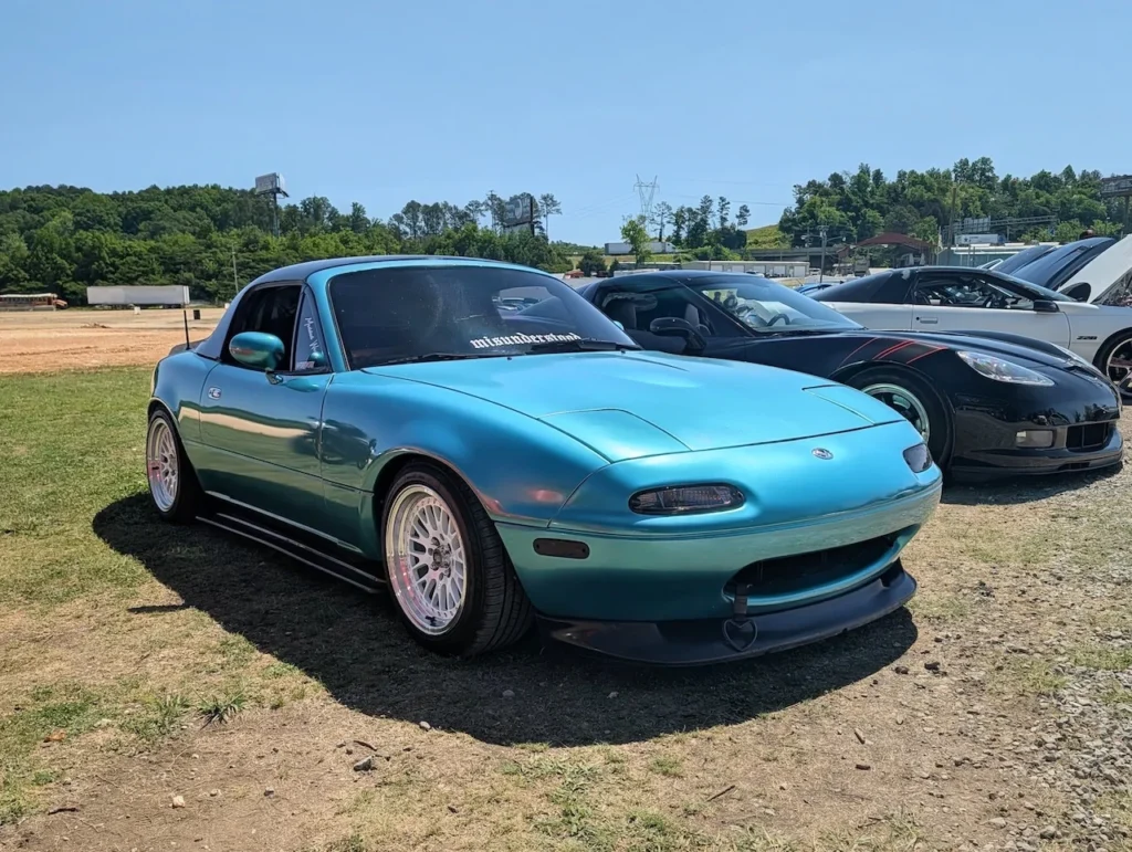 A turquoise Mazda MX-5 Miata parked on grass with another car in the background under clear sky.