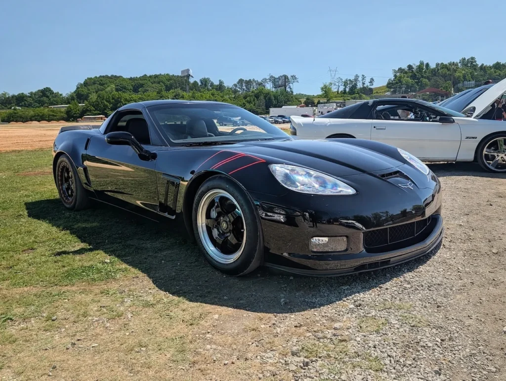 Black Chevrolet Corvette parked on grass at a car event with another vehicle visible behind it.