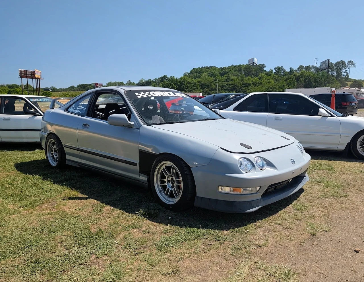 Silver Acura Integra parked on grass, with other vehicles in the background under a clear blue sky.