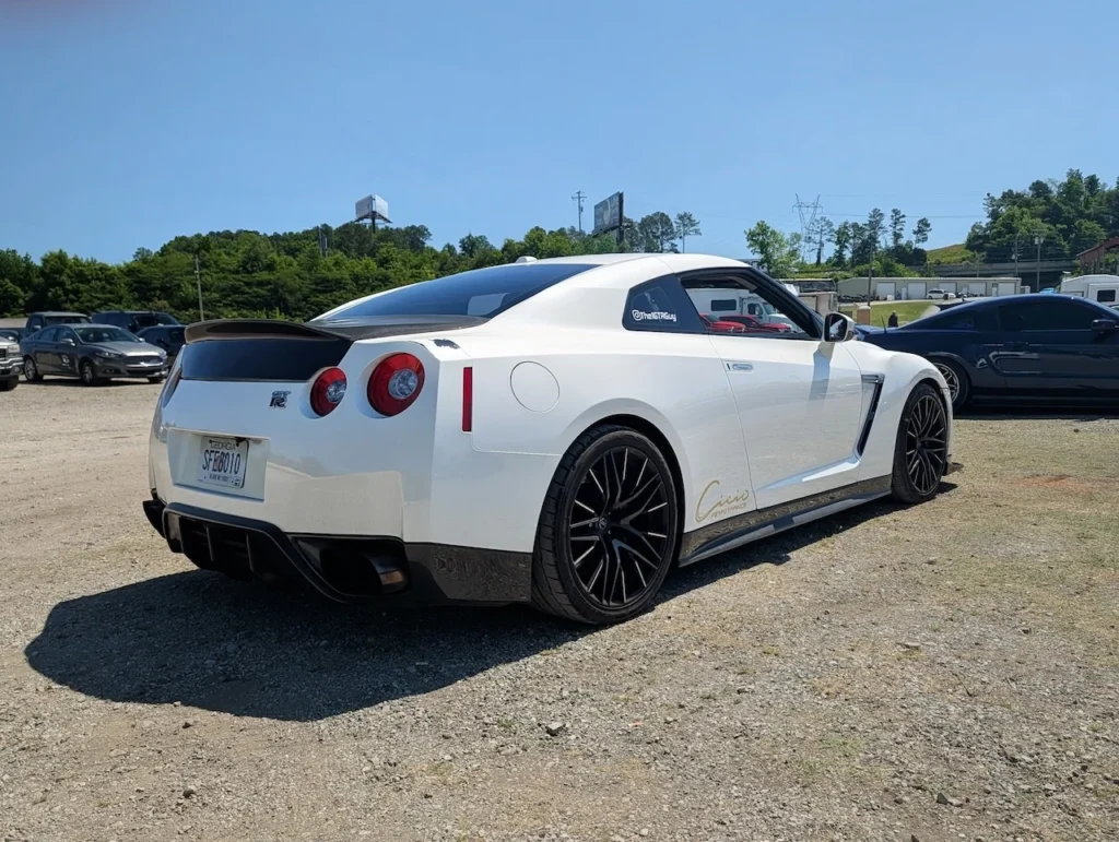 White Nissan GT-R parked on gravel with other vehicles in the background and blue sky above.
