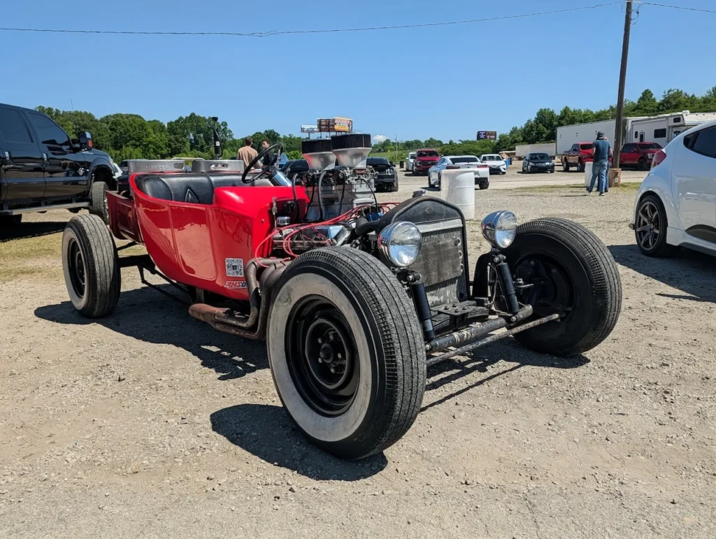 Red modified vintage hot rod with no roof in a parking area on a sunny day.