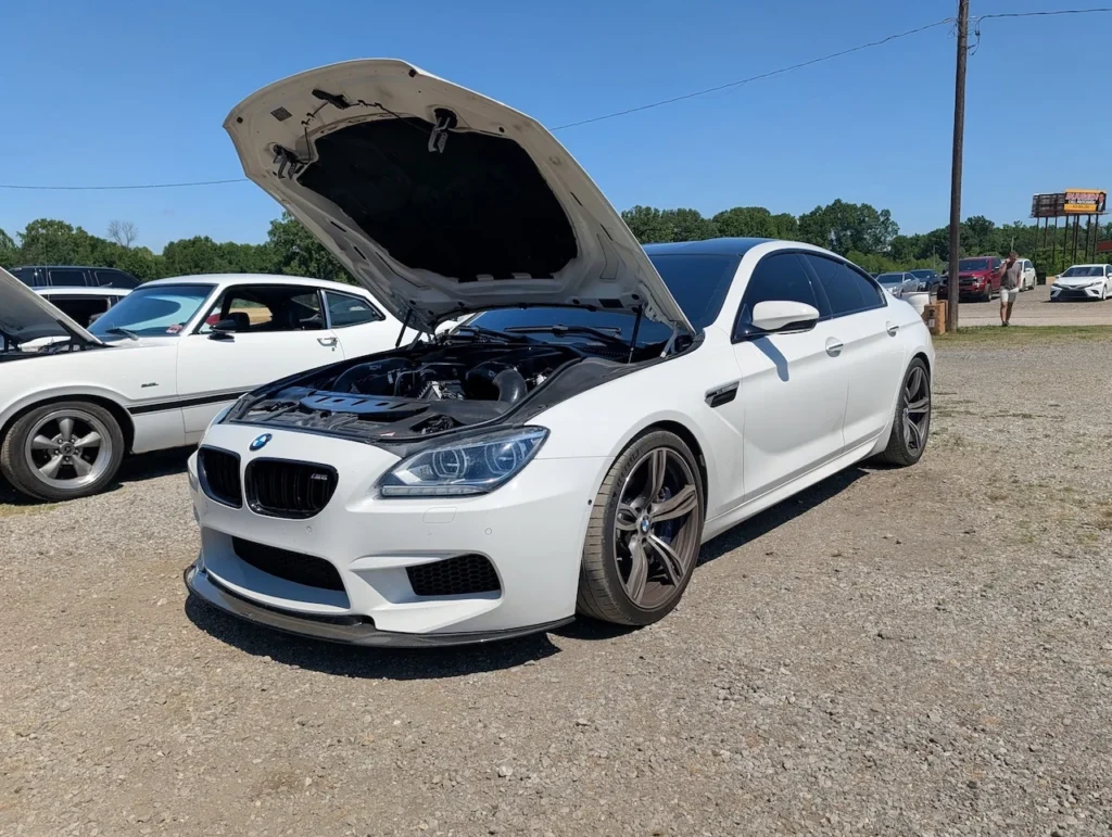 White BMW M6 with hood raised, showing engine in a gravel area alongside other vehicles.