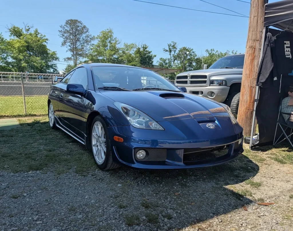 Blue Toyota Celica parked on grass with a truck and tents in the background.