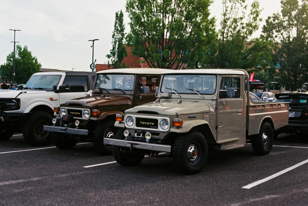 Tan and brown classic Toyota Land Cruisers and a white Ford Bronco parked in lot with trees and people in background