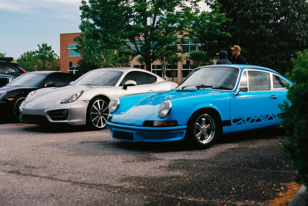 Blue Porsche 911 Carrera parked next to a silver Porsche Cayman in a parking lot with trees and a building in the background