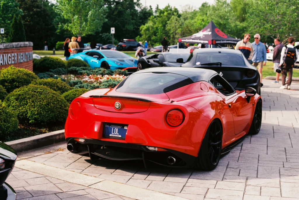 Red Alfa Romeo 4C parked with a blue Lamborghini Huracán and black Ferrari in a green outdoor car event setting