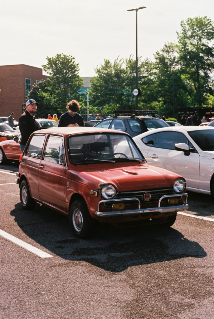 Rust-colored Honda N600 hatchback parked in a lot with people and other vehicles around during a sunny day