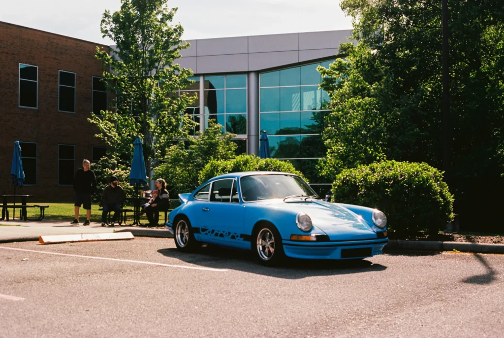 Light blue Porsche 911 parked in front of a modern building with trees and people sitting at picnic tables.