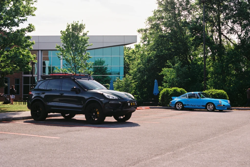 Black Porsche Cayenne SUV and blue Porsche 911 coupe parked in a lot with trees and modern building in background