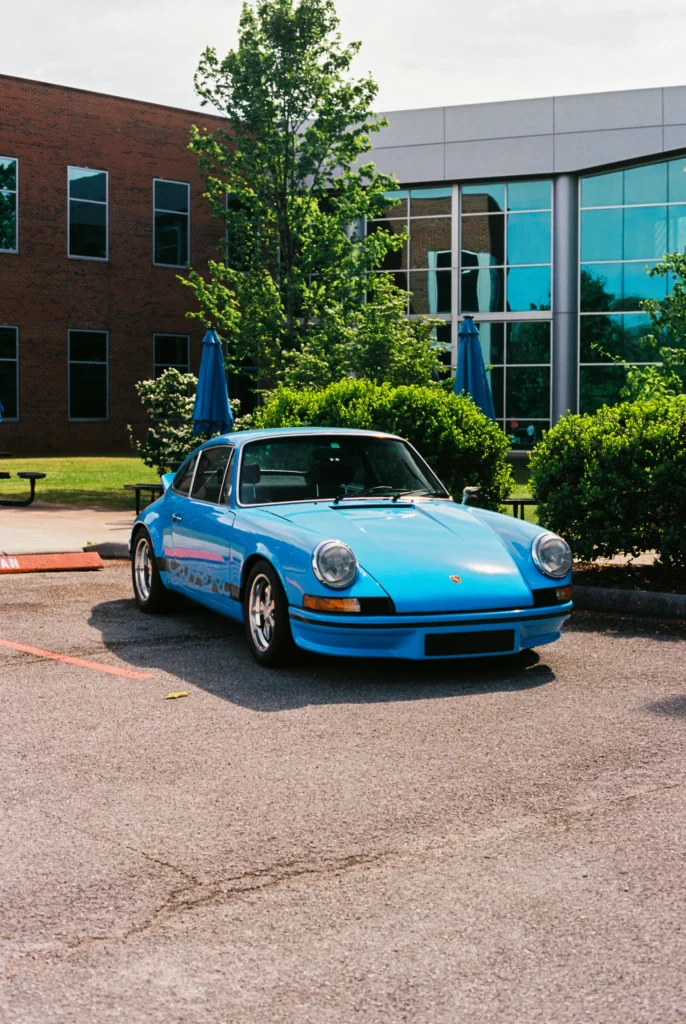 Blue Porsche parked in front of brick and glass building with greenery and blue umbrellas