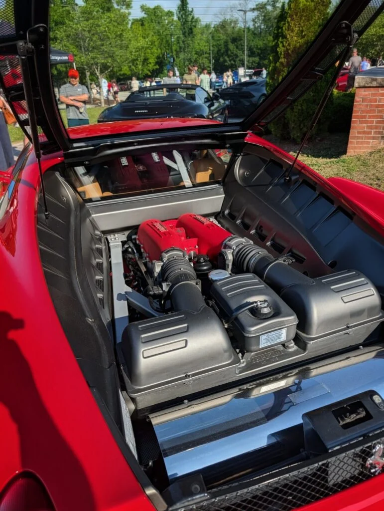 Red Ferrari F430 engine bay with red intake manifolds and visible crowd in background at outdoor car event