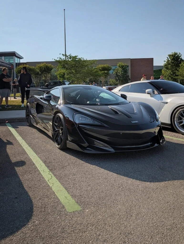 Black McLaren 600LT parked next to a white Nissan GT-R at an outdoor car event with people and trees in background.