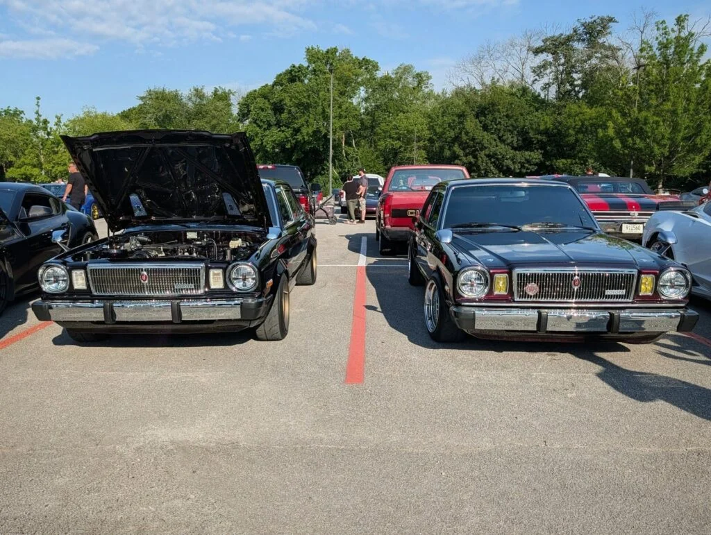 Two Toyota Cressidas parked in a lot at a car meet, one with the hood open showing the engine bay, surrounded by other vehic