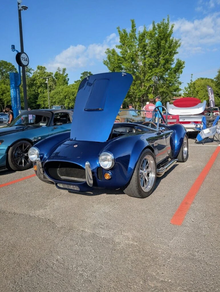 Dark blue Shelby Cobra with hood open parked beside a blue convertible at a car meet on a sunny day