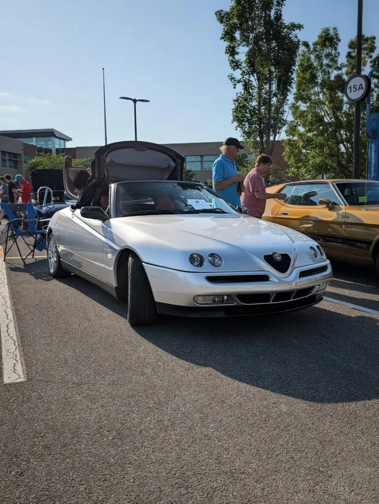 Silver Alfa Romeo convertible with roof partially raised parked outdoors at a car event with two men standing beside a yellow