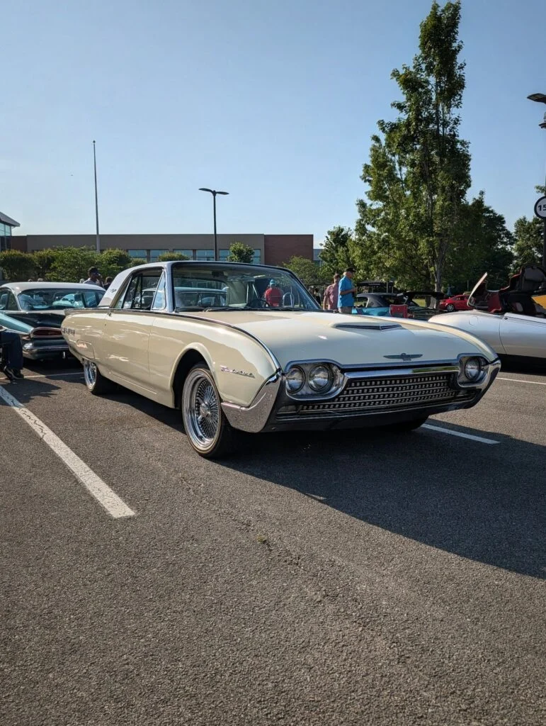 White Ford Thunderbird parked at an outdoor car event with other cars and people in the background.