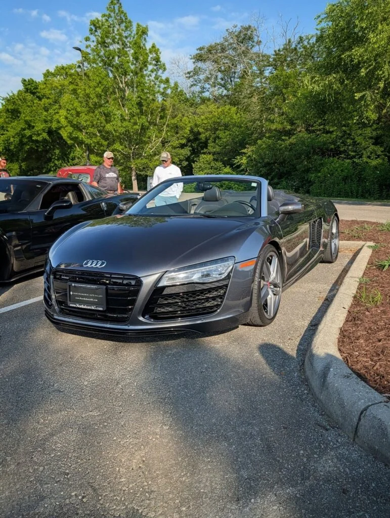 Black Audi R8 Spyder convertible parked in a lot with two men standing nearby and trees in the background