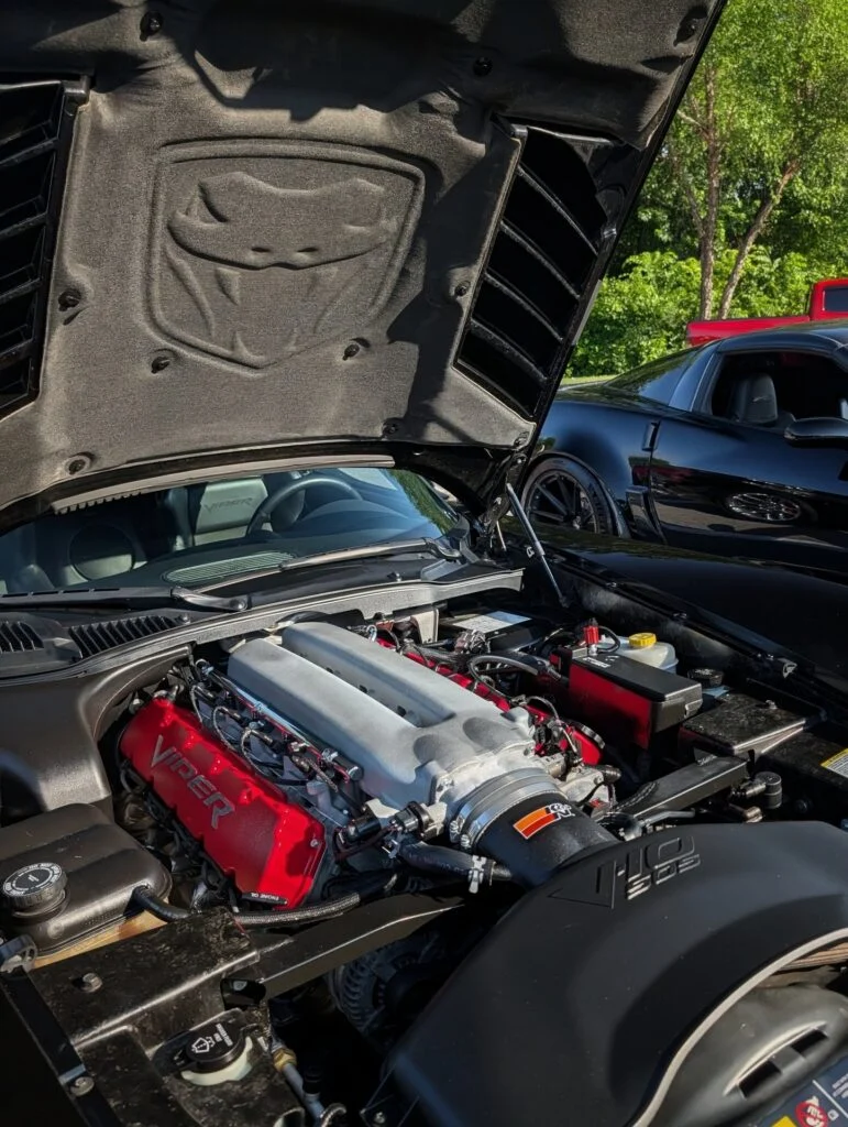 Open hood showing engine bay with bright red Viper valve covers and black intake on black Dodge Viper at outdoor car event