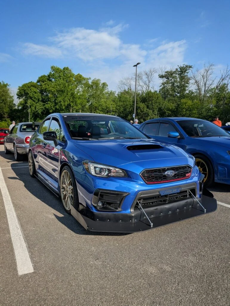 Blue Subaru WRX STI with a front splitter parked in a lot with other cars and trees in the background