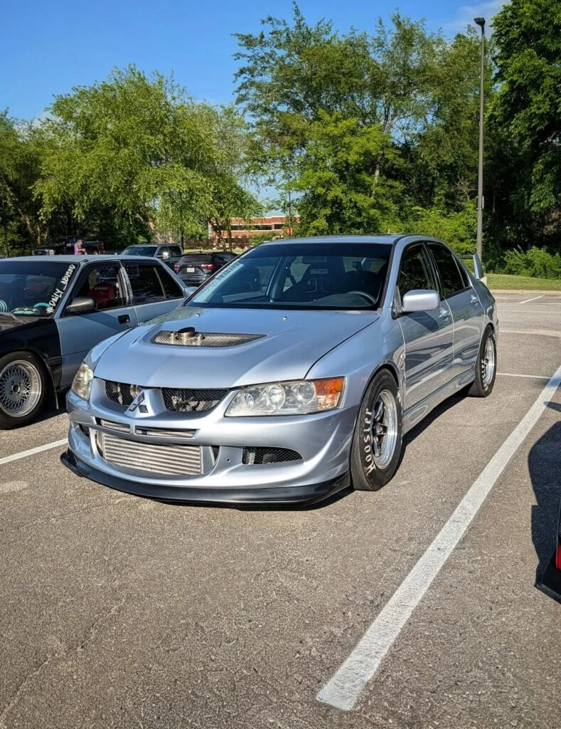 Silver Mitsubishi Lancer Evolution parked in a lot with other cars and green trees in the background.
