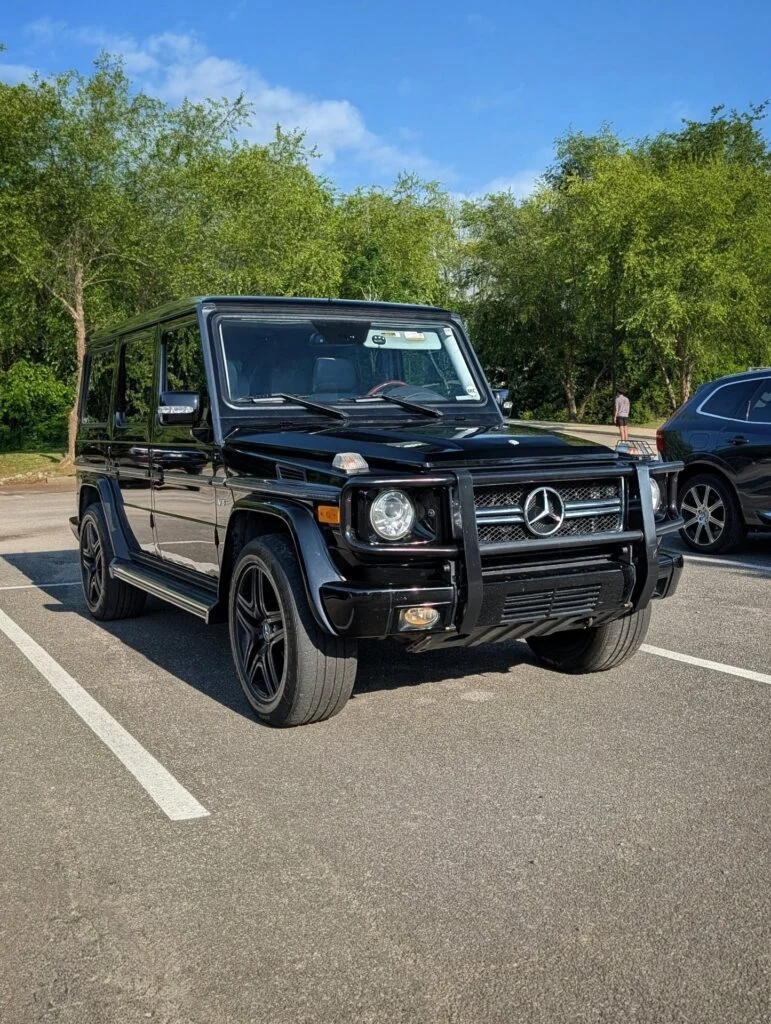 Black Mercedes-Benz G-Class SUV parked in an outdoor lot with trees and a clear blue sky in the background