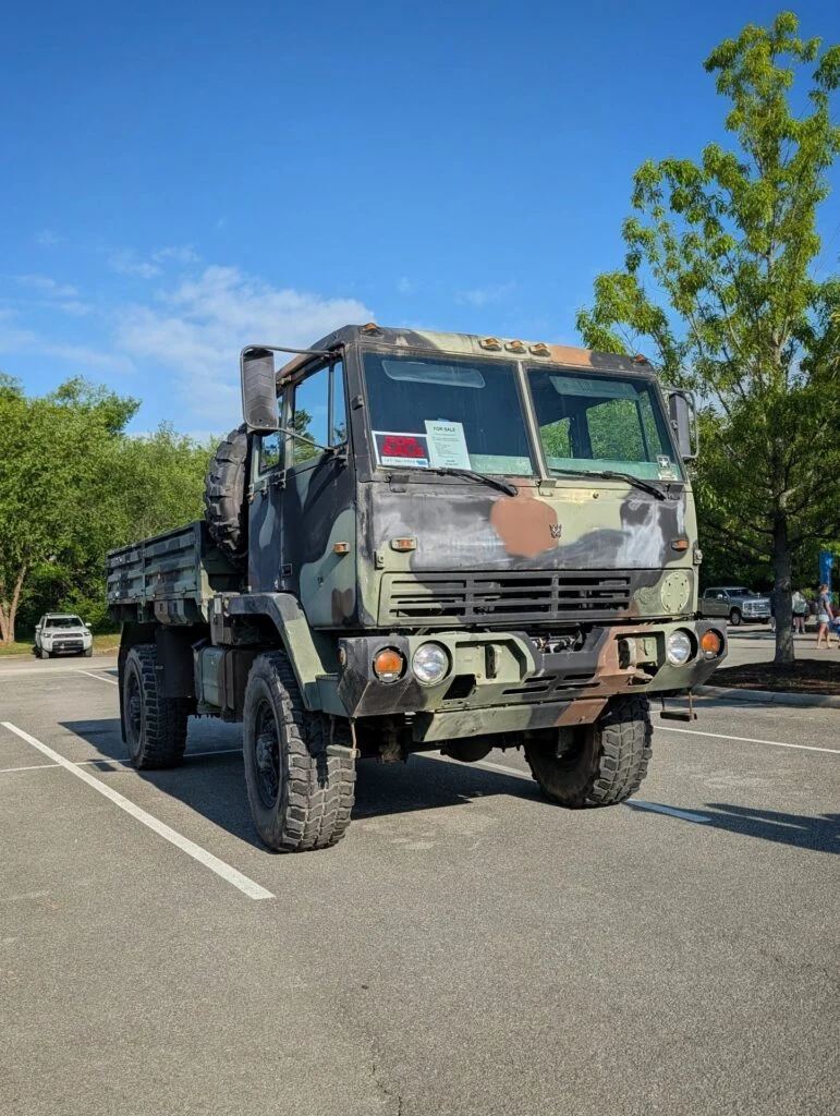 Military-style camo-painted 6x6 truck parked in lot with trees and blue sky background