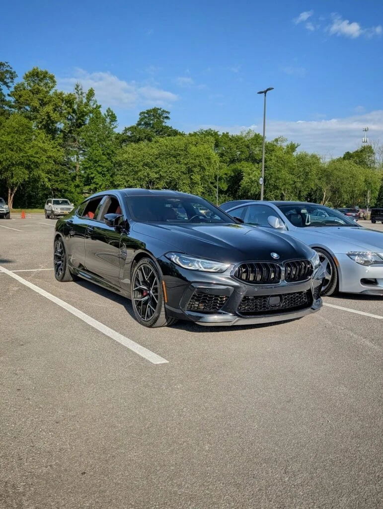 Black BMW M8 sedan parked in a lot with other vehicles under a partly cloudy blue sky.