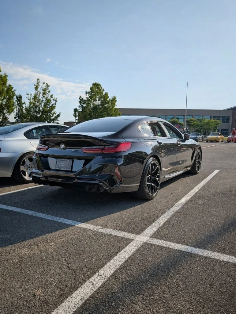Black BMW M8 sedan parked in an outdoor lot with other cars and trees in background