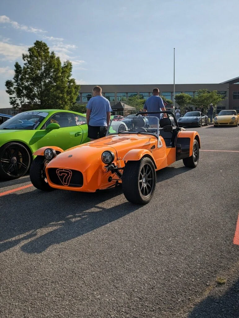 Orange Caterham Seven roadster parked next to a green Porsche 911 in an outdoor parking lot with people and cars in the backg